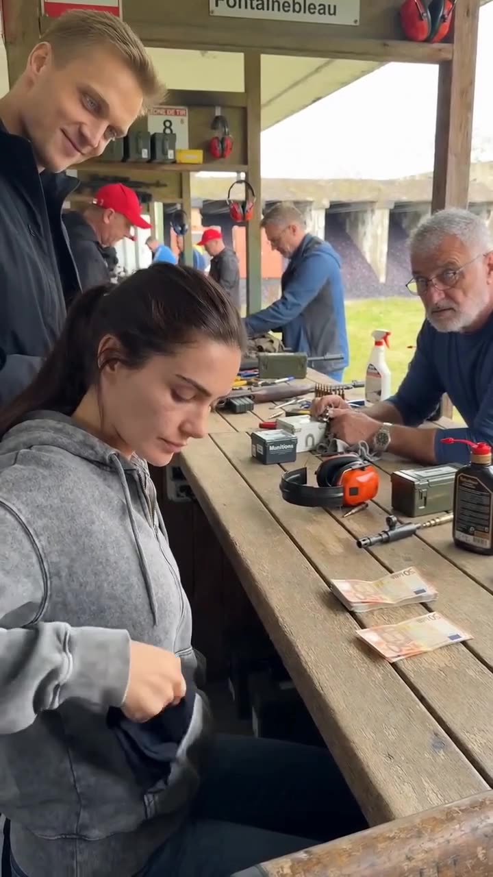 Stacks of euro cash sit on the shooting bench while the woman quietly prepares a black blindfold.
