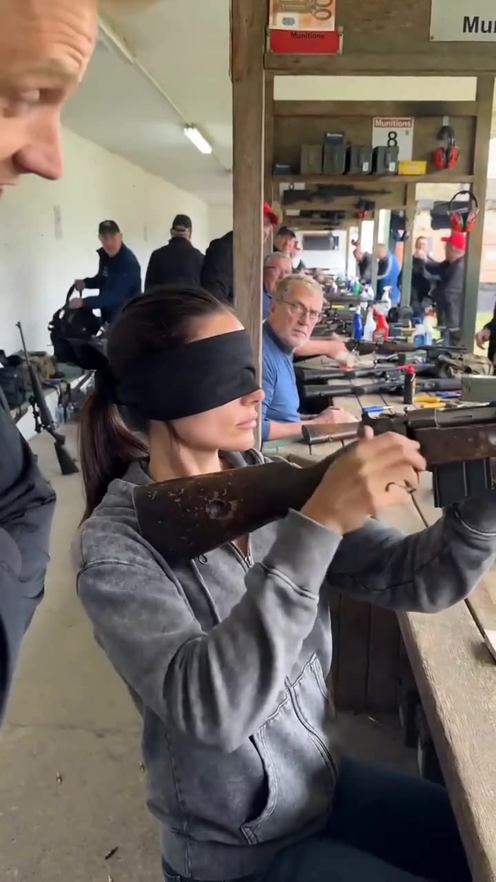 Blindfolded, the woman raises the rifle to take aim, astounding the onlookers around her.