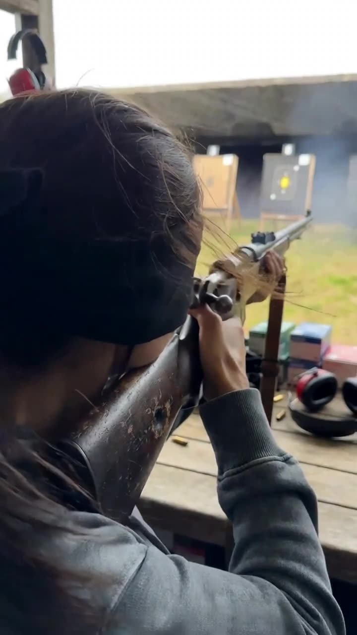 Over-the-shoulder view of Camille firing the rifle downrange toward paper targets