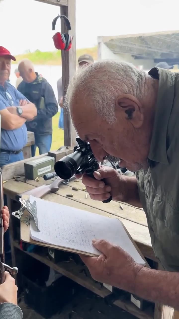 A senior range official holds a clipboard while peering through a scope at the shooting range.