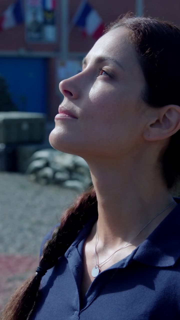 Camille tilts her chin upward, revealing a hidden silver chain and metallic insignia.