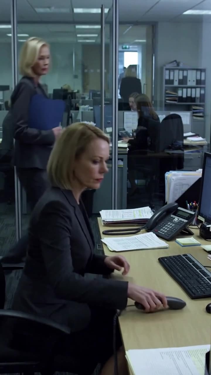 Evelyn sets down the desk phone inside her glass office as colleagues work in the background.