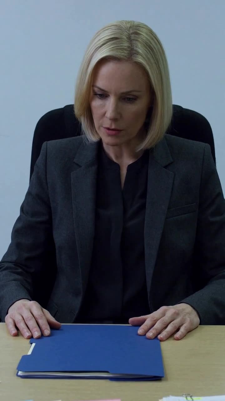 Evelyn sits stone-faced in her grey blazer, staring down skeptically at the unopened folder.