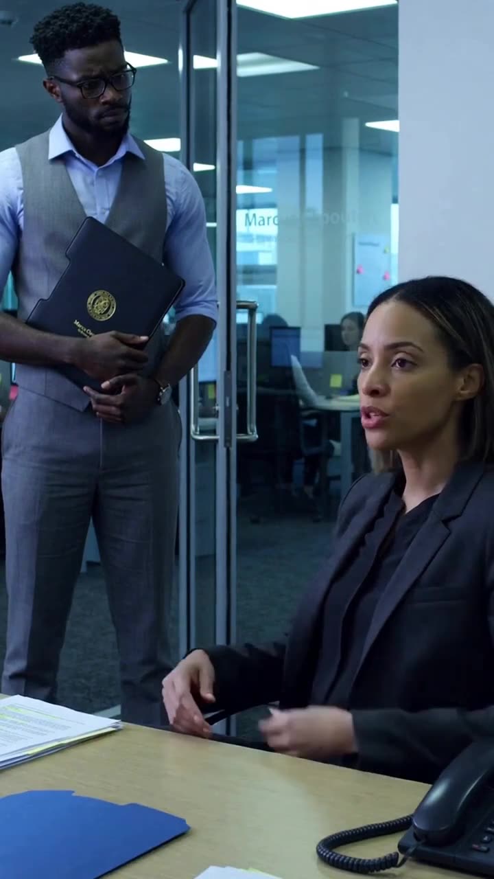 Female manager speaking decisively across the desk with a man holding a portfolio in the background