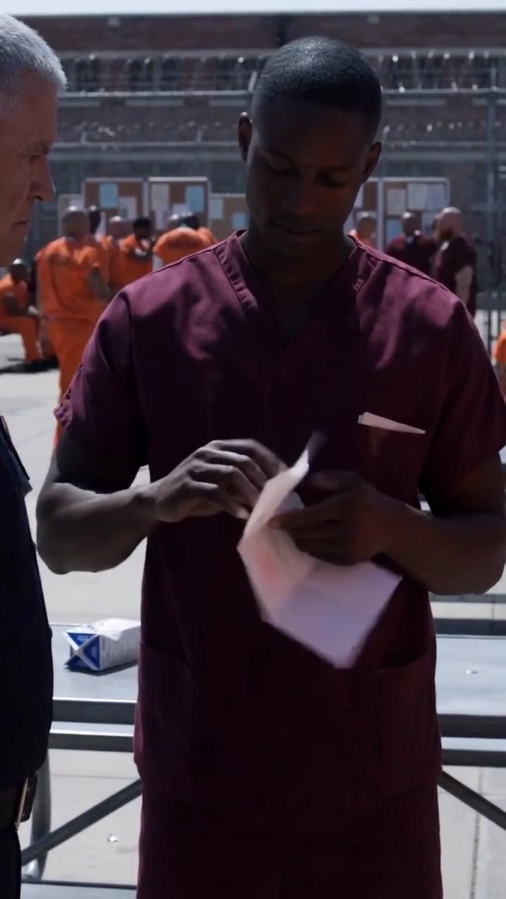 The inmate in burgundy scrubs unfolds official documents before the corrections officer.