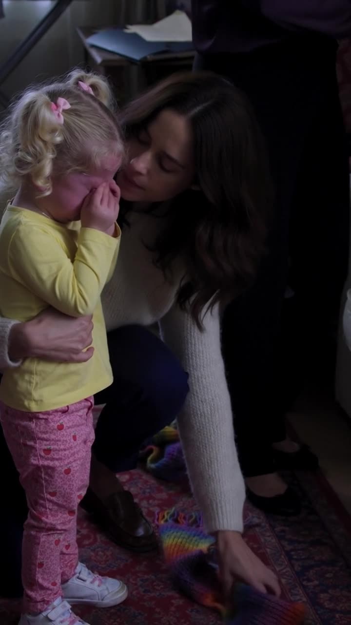 Camille kneels down to comfort Léa and retrieve the knitted rainbow scarf