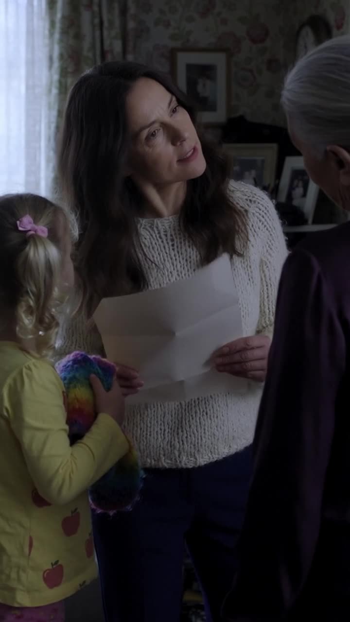 Camille holds a folded white document while facing a sceptical Monique and young Léa.