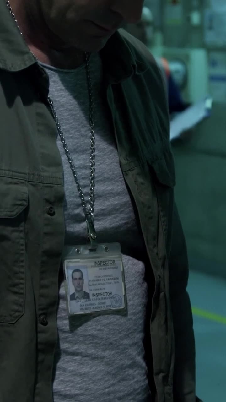 A close-up view of the steel inspector credential hanging against Marc's olive jacket.