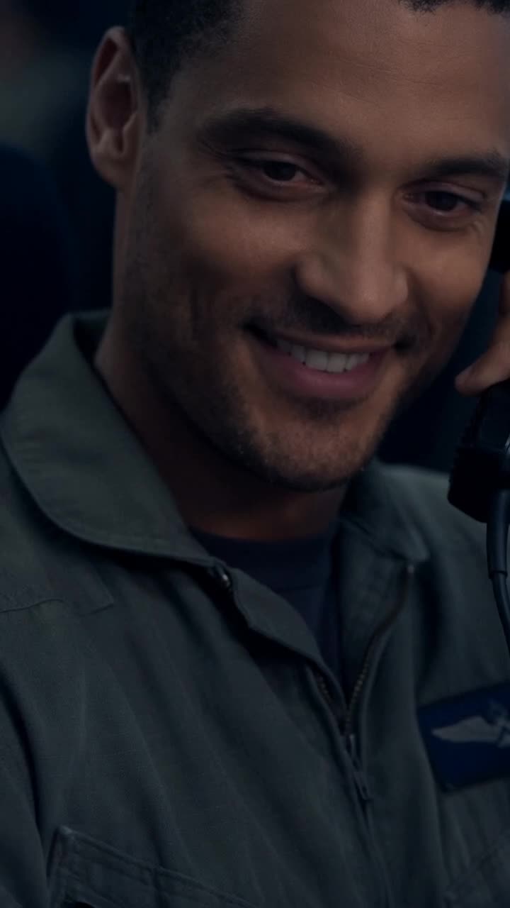 Close-up of Julien wearing a military jumpsuit while smiling over the phone.
