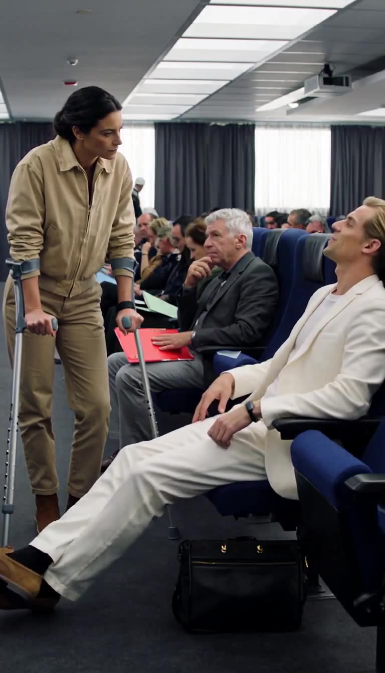 Julien reclining sarcastically in a white suit with his legs outstretched across Camille's path